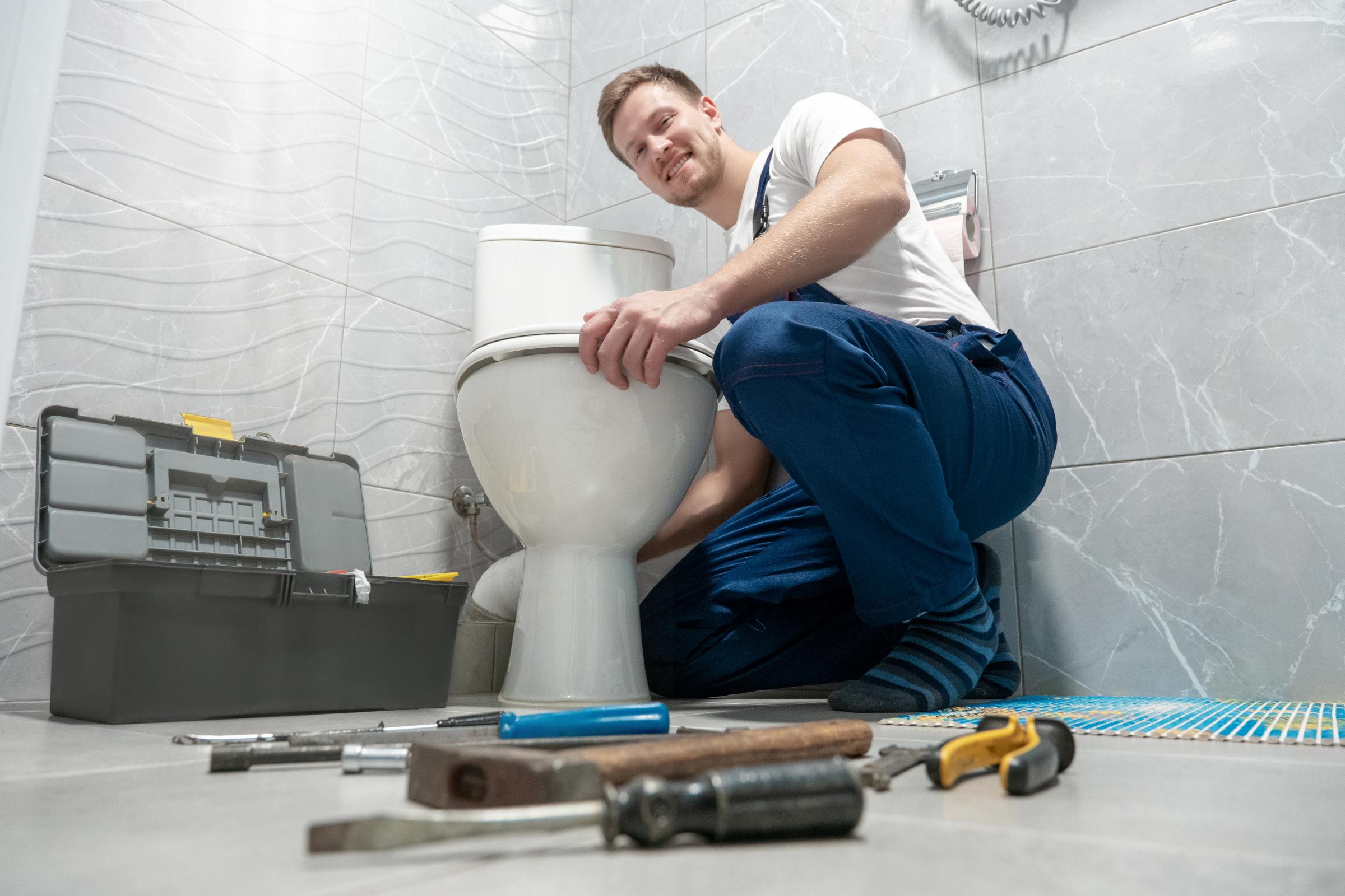 smiling man plumber in uniform repairing toilet bowl using instrument kit looks happy professional repair service..